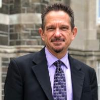 Dr. Kirk Bingaman wears a suit while standing in front of a grey stone building.